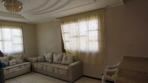 A living room with a sectional sofa, a small white chair, and a ceiling light fixture. - Villa 400m2 with swimming pool 10 min from the beach tifnit (Agadir)
