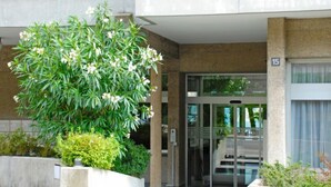 Une entrée d’immeuble avec une porte en verre, un bac à fleurs en béton contenant un arbre et une plaque portant le numéro « 15 ».