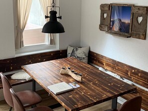 Een houten eettafel met een zwart hanglamp, een bank met kussen en een aan de muur hangend fotolijstje.
