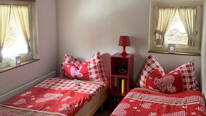 Une chambre avec deux lits, chacun recouvert d’une couette à carreaux rouges et blancs, une lampe rouge posée sur une table de chevet et une fenêtre avec des rideaux.