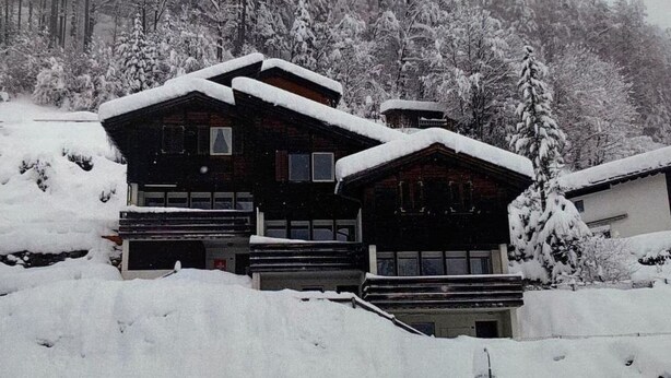Un grand chalet en bois doté de nombreux balcons, entouré d’arbres couverts de neige et situé au pied d’une pente abrupte.