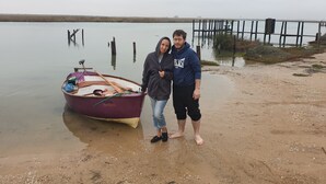 Dos personas de pie en una orilla arenosa cerca de un bote con remos y una tabla de remo.