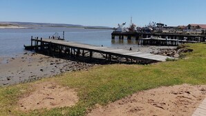Una zona costera con un muelle de madera que se adentra en aguas tranquilas, una playa de césped y una costa lejana.