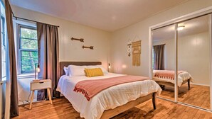 A bedroom with a wooden floor, a bed with white bedding and a yellow pillow, a bedside table, a mirror, and a window with curtains. - Centrally Located Hot Springs Home w/ Deck! (Hot Springs)