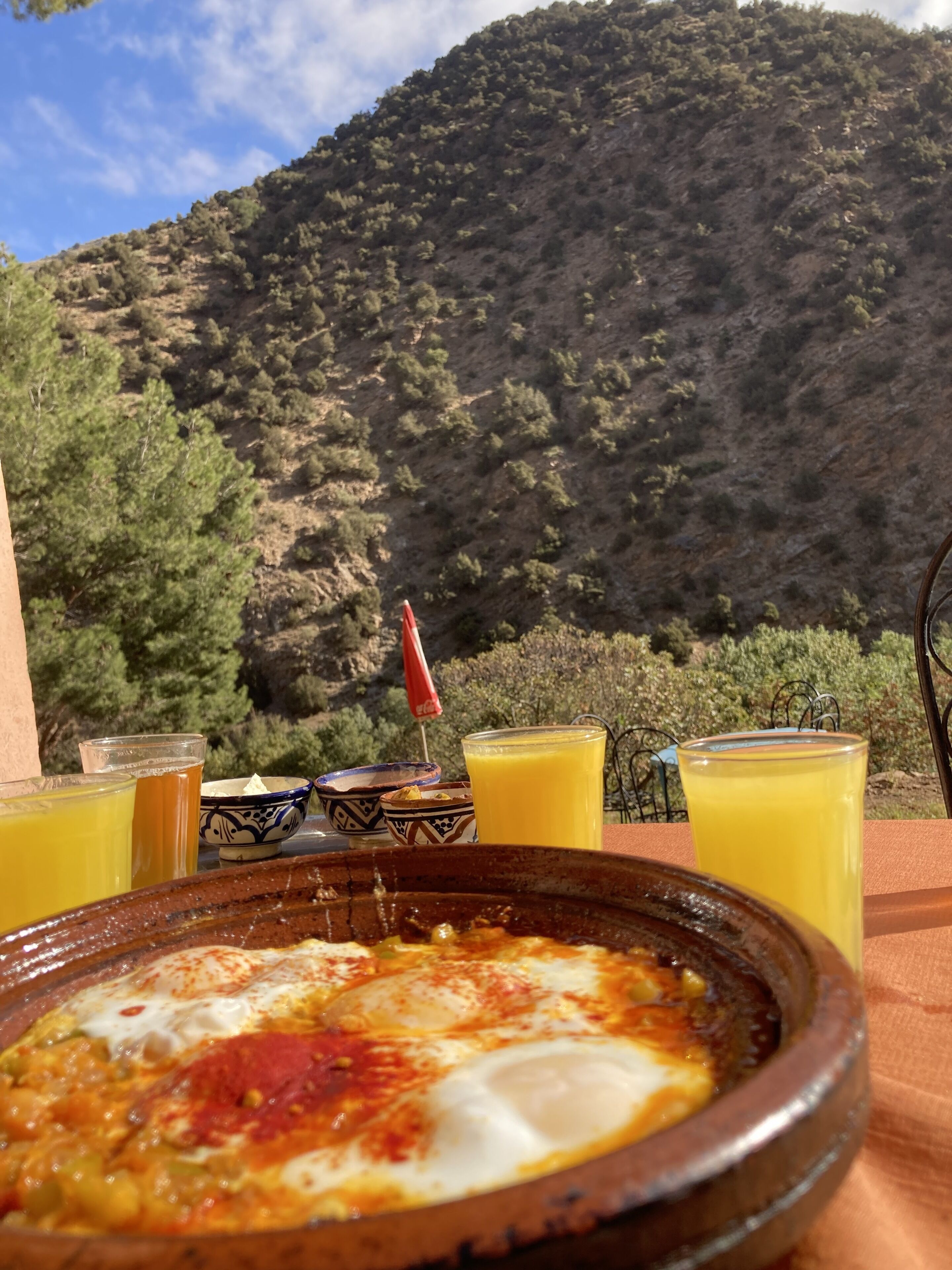 Una mesa con un cuenco de comida, dos vasos de zumo de naranja y un paraguas rojo. El fondo muestra un paisaje montañoso.