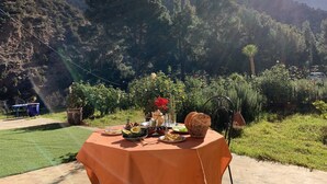 Mesa de comedor al aire libre con comida y bebidas, rodeada de vegetación exuberante y un paisaje montañoso.