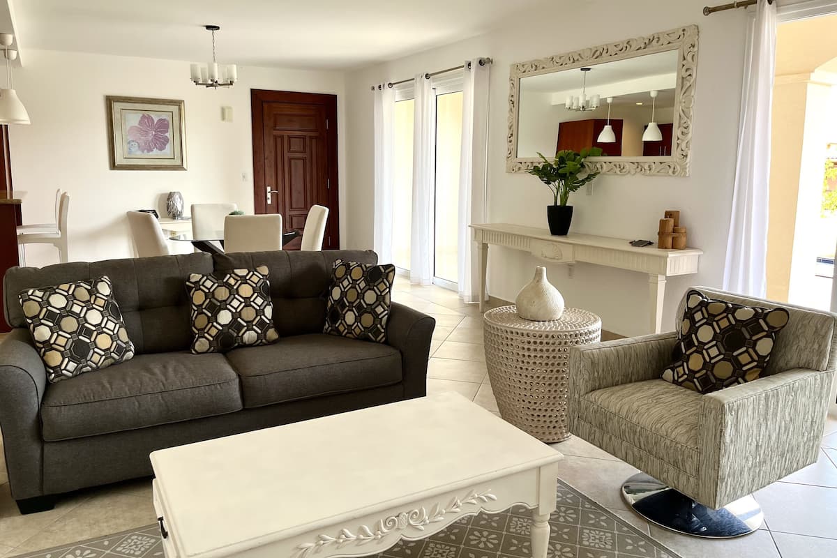 Ein Wohnzimmer mit einem dunkelgrauen Sofa, einem weißen Couchtisch und einem gemusterten Sessel. An einer Konsole stehen ein großer Spiegel und eine Vase mit Blumen.