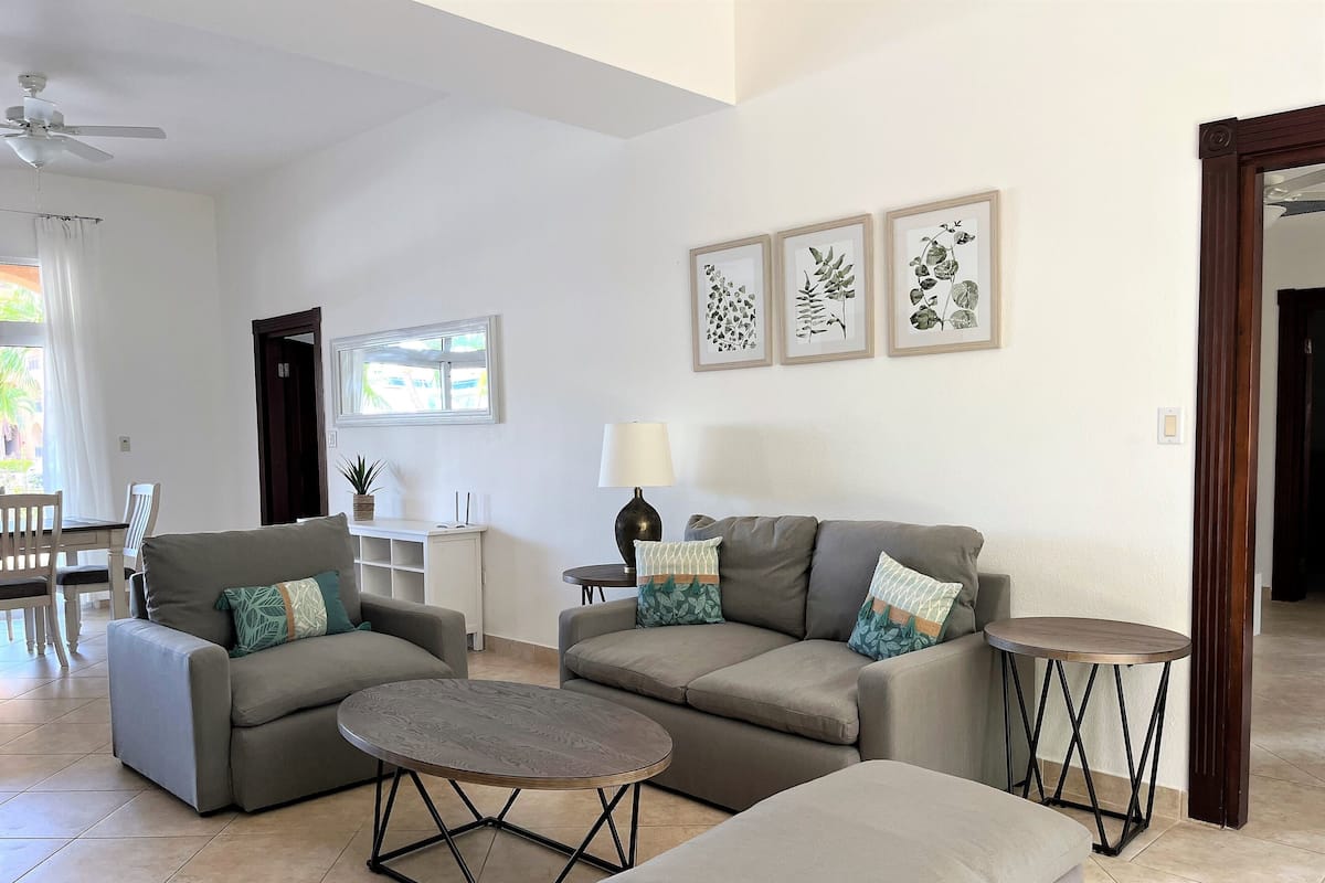 Ein modernes Wohnzimmer mit einer grauen Sofagarnitur, einem runden Couchtisch und einem kleinen Beistelltisch. Ein Fenster mit Vorhängen, ein Deckenventilator und gerahmte Pflanzenmotive schmücken die Wand.
