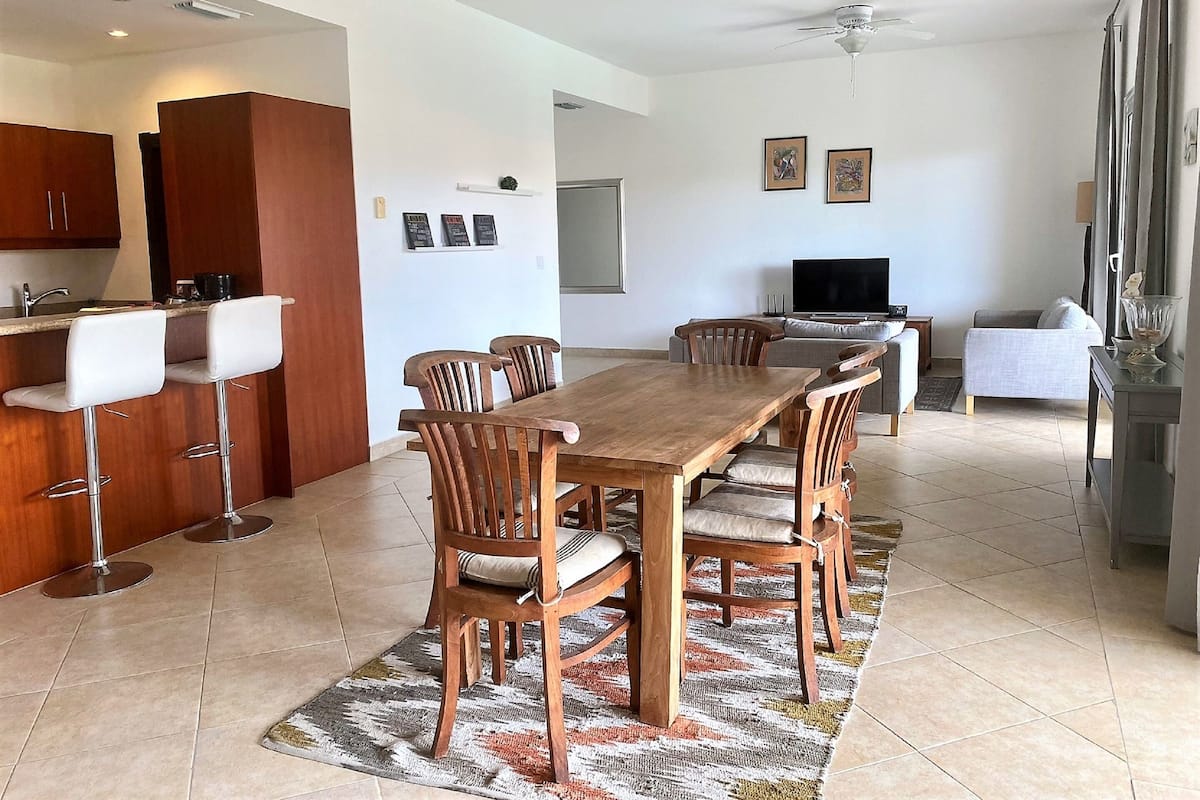 Eine moderne Küche mit Holzausstattung, ein Essbereich mit Holztisch und -stühlen, eine Bar mit weißen Hockern, ein graues Sofa und ein Fernseher im Wohnzimmer.