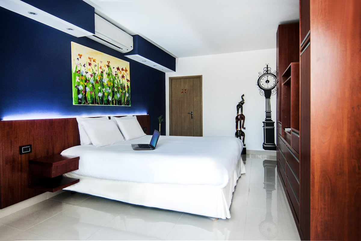 A modern hotel room with a large bed, a laptop on the bed, a wooden wardrobe, a clock, and a painting on the wall.