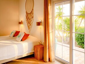 A bedroom with a bed, two wall-mounted lamps, a decorative wall hanging, a small round table, and a large window with a view of greenery.