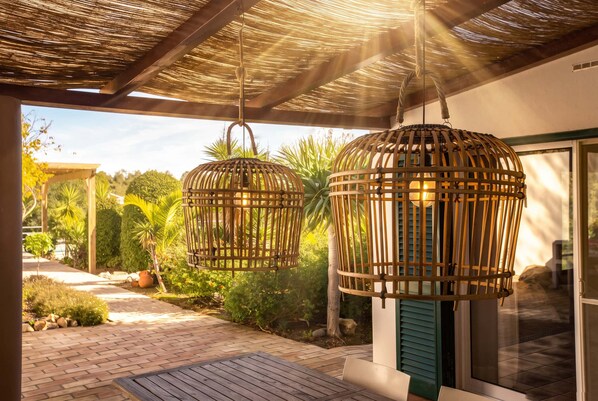 A covered outdoor area with two hanging wicker lamps, a brick pathway, and lush greenery.