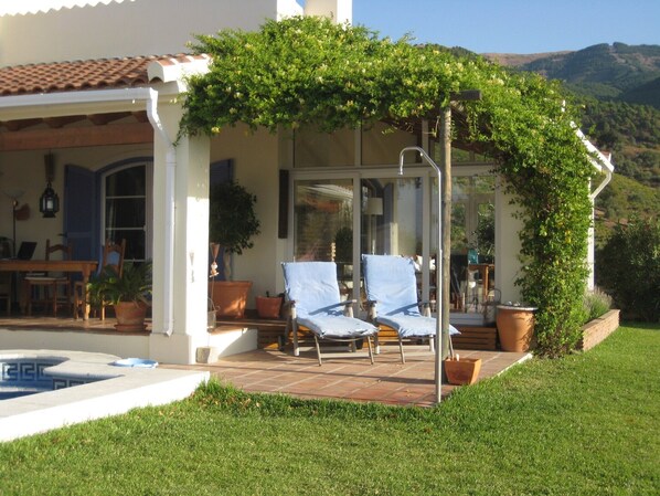 A covered outdoor seating area with two lounge chairs, a glass door entrance, and a patio with potted plants.