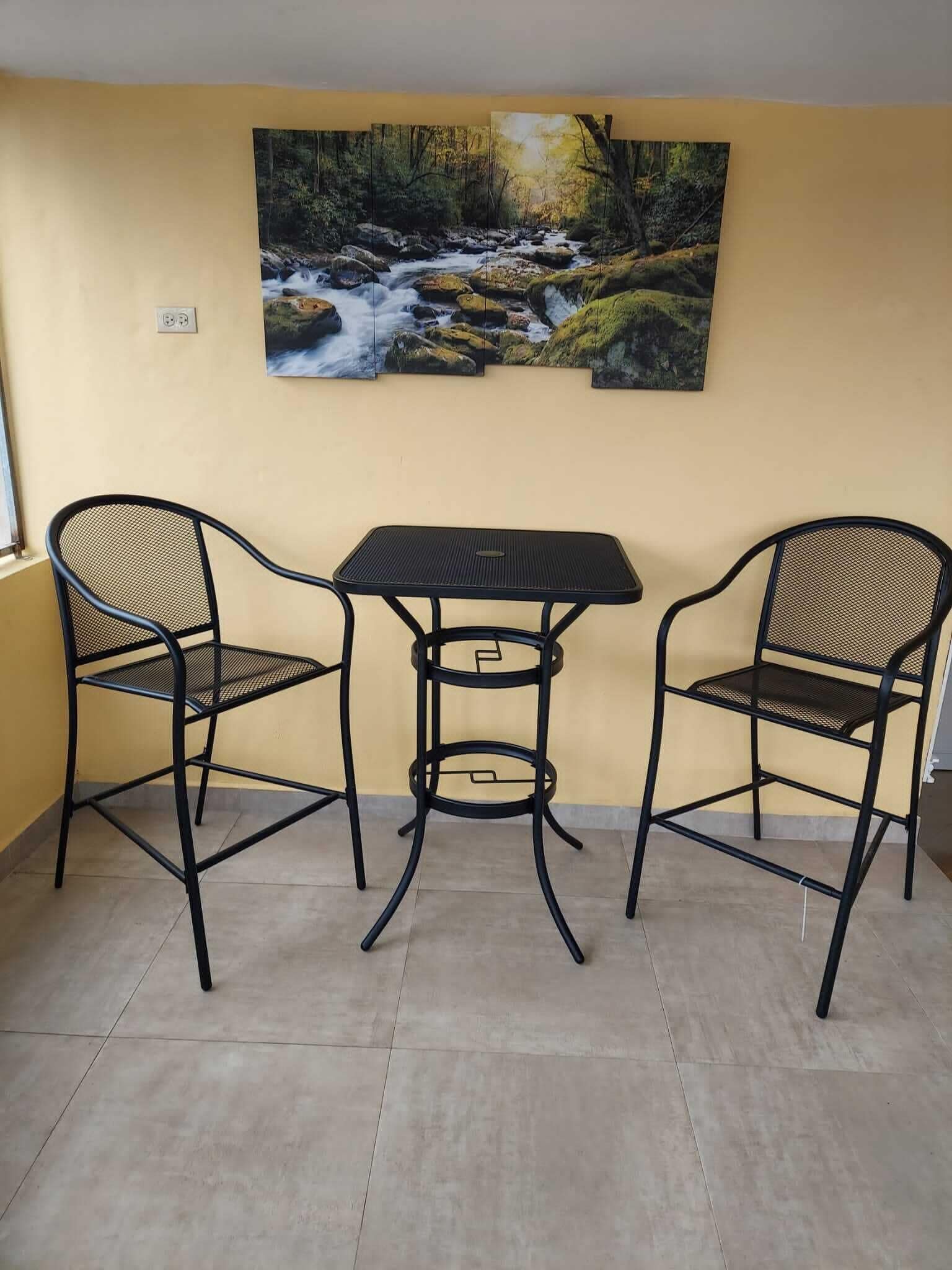 Een kamer met een gele wand, een schilderij van een bosbeek, en een metalen tafel met twee stoelen.