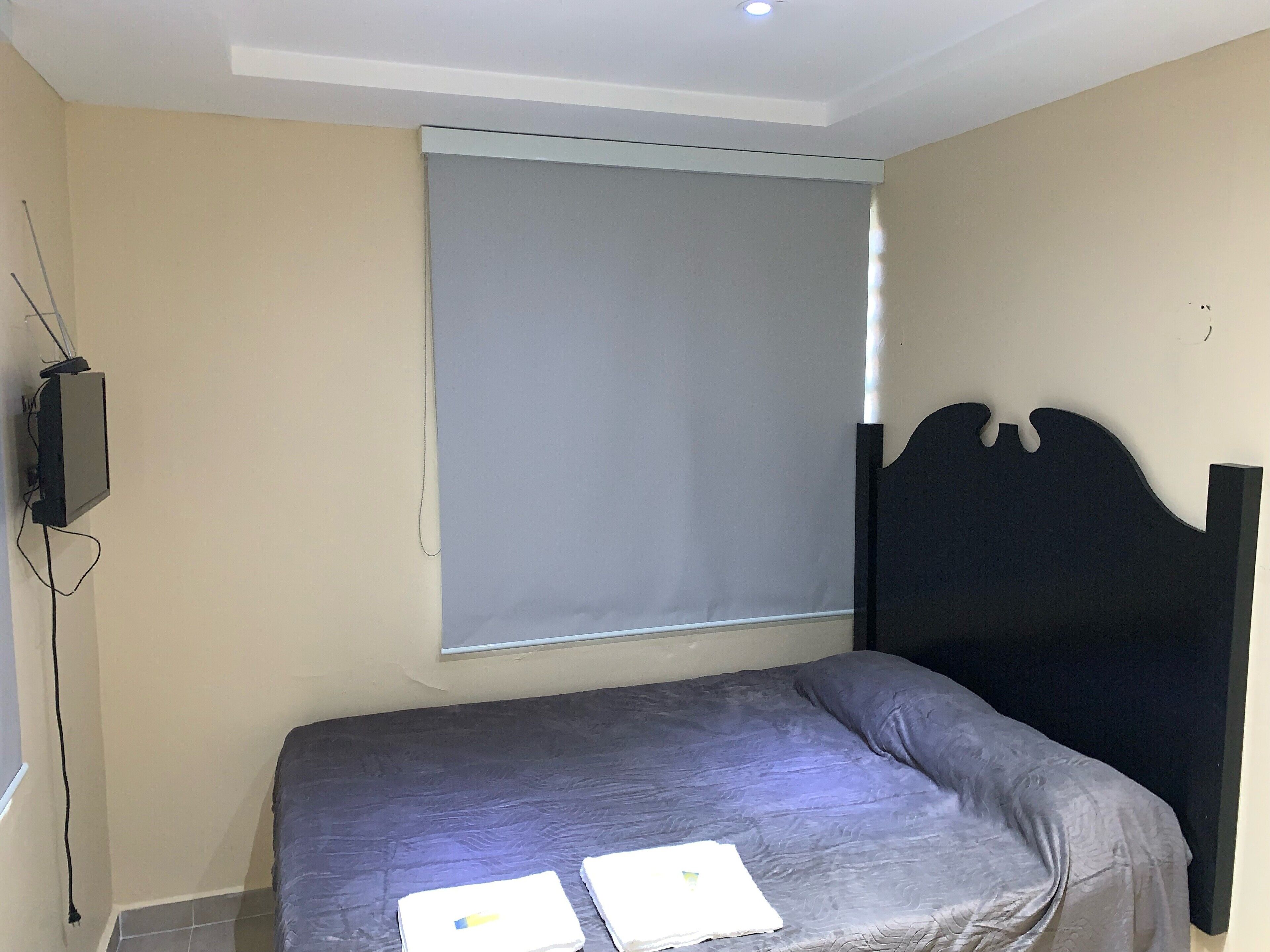 A small bedroom with a single bed, a window with a blind, a television mounted on the wall, and a power outlet.