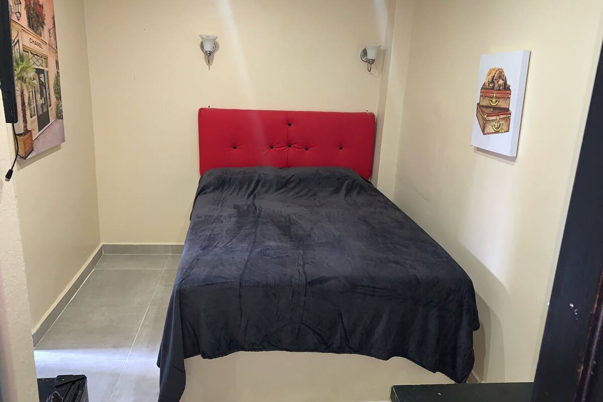A small, neatly arranged bedroom with a red headboard, a black bedspread, and a single wall-mounted light fixture.