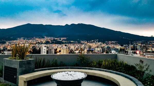 Paisagem urbana com montanha ao fundo, uma ampla área circular com assentos e uma lareira, além de vista para as luzes da cidade.