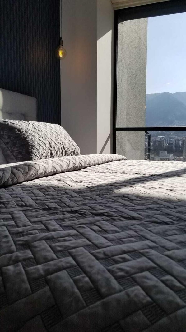Una cama con cubrecama acolchado, una lámpara colgante y vista a un paisaje urbano a través de un ventanal grande.