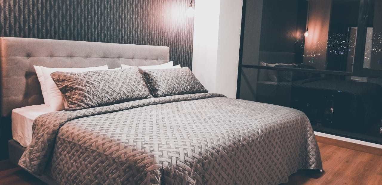 A neatly made bed with patterned bedding, a tufted headboard, and a view of the city lights through a large window.