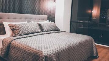 A neatly made bed with patterned bedding, a tufted headboard, and a view of the city lights through a large window.