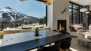 A modern interior with a large dining table, a fireplace, and a view of a snow-capped mountain. - Lone Peak Views | Hot Tub | Ski-In/Out (Big Sky)