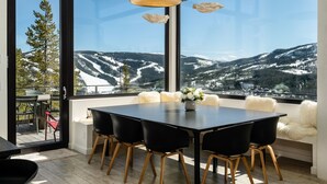 A dining area with a table and chairs, large windows with a mountain view, and hanging pendant lights. - Lone Peak Views | Hot Tub | Ski-In/Out (Big Sky)