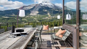 A wooden deck with a table set for dining, chairs, and a glass railing overlooking a mountainous landscape. - Lone Peak Views | Hot Tub | Ski-In/Out (Big Sky)
