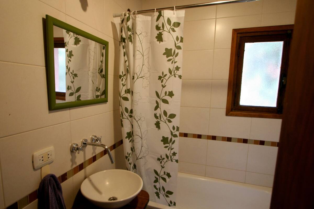 Ein Badezimmer mit einem weißen Waschbecken, einem Spiegel mit grünem Rahmen, einem Duschvorhang mit Blumenmuster und einem Fenster über der Badewanne.