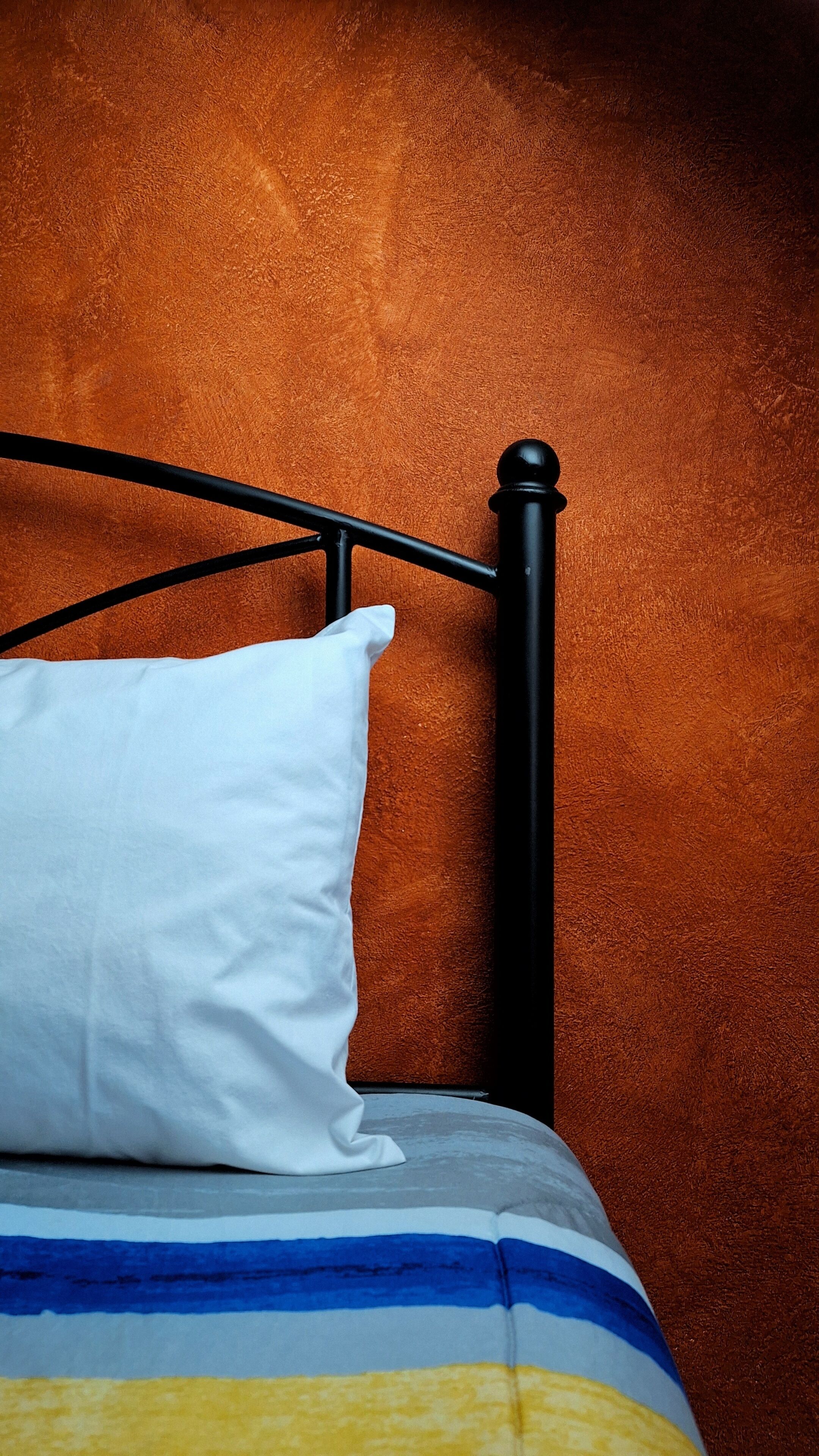 Una cama con un almohadón blanco y una manta a rayas frente a una pared naranja con textura.