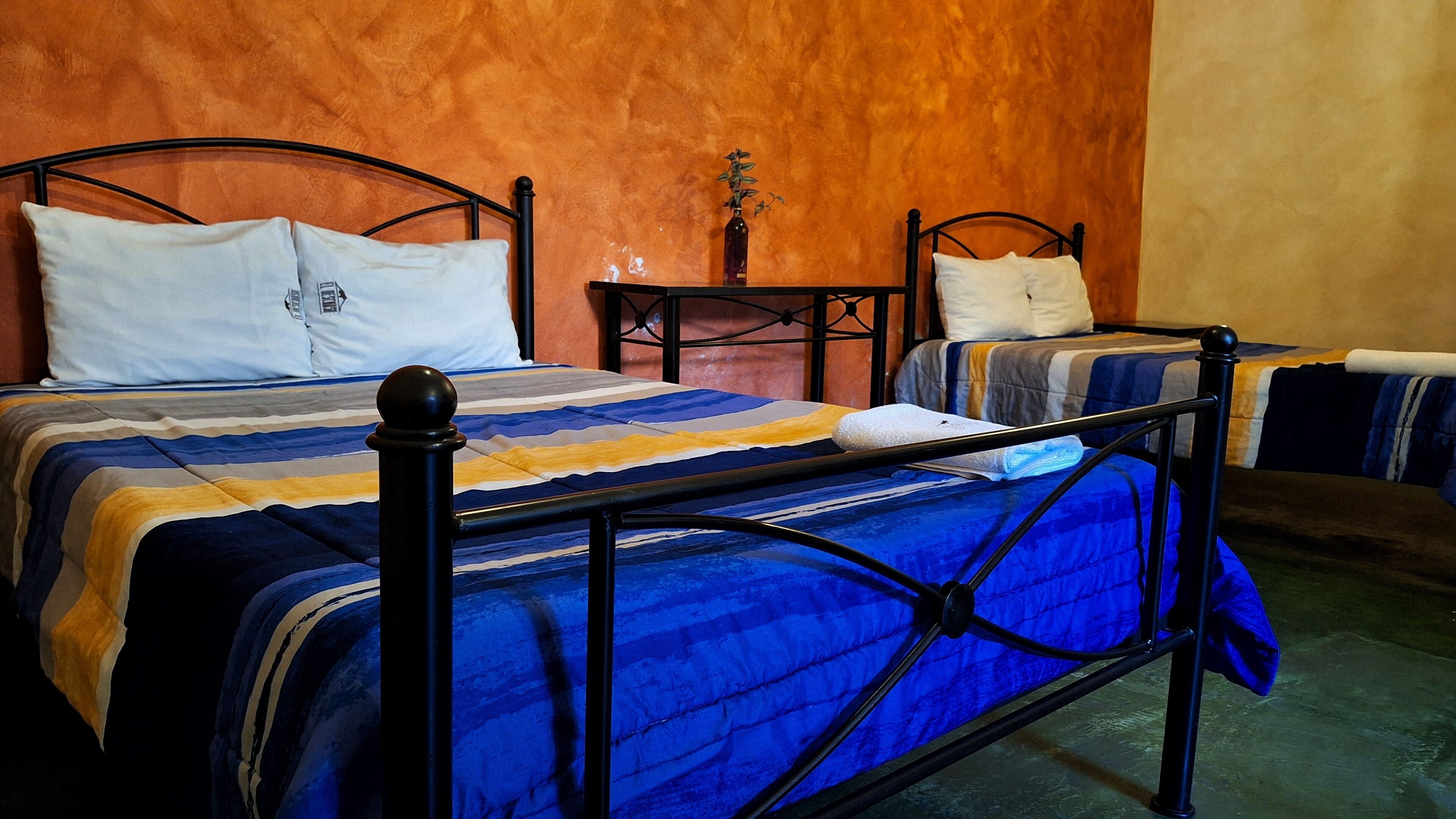 A room with two beds, each with a black metal frame and blue bedding. There is a small table with a vase between the beds.
