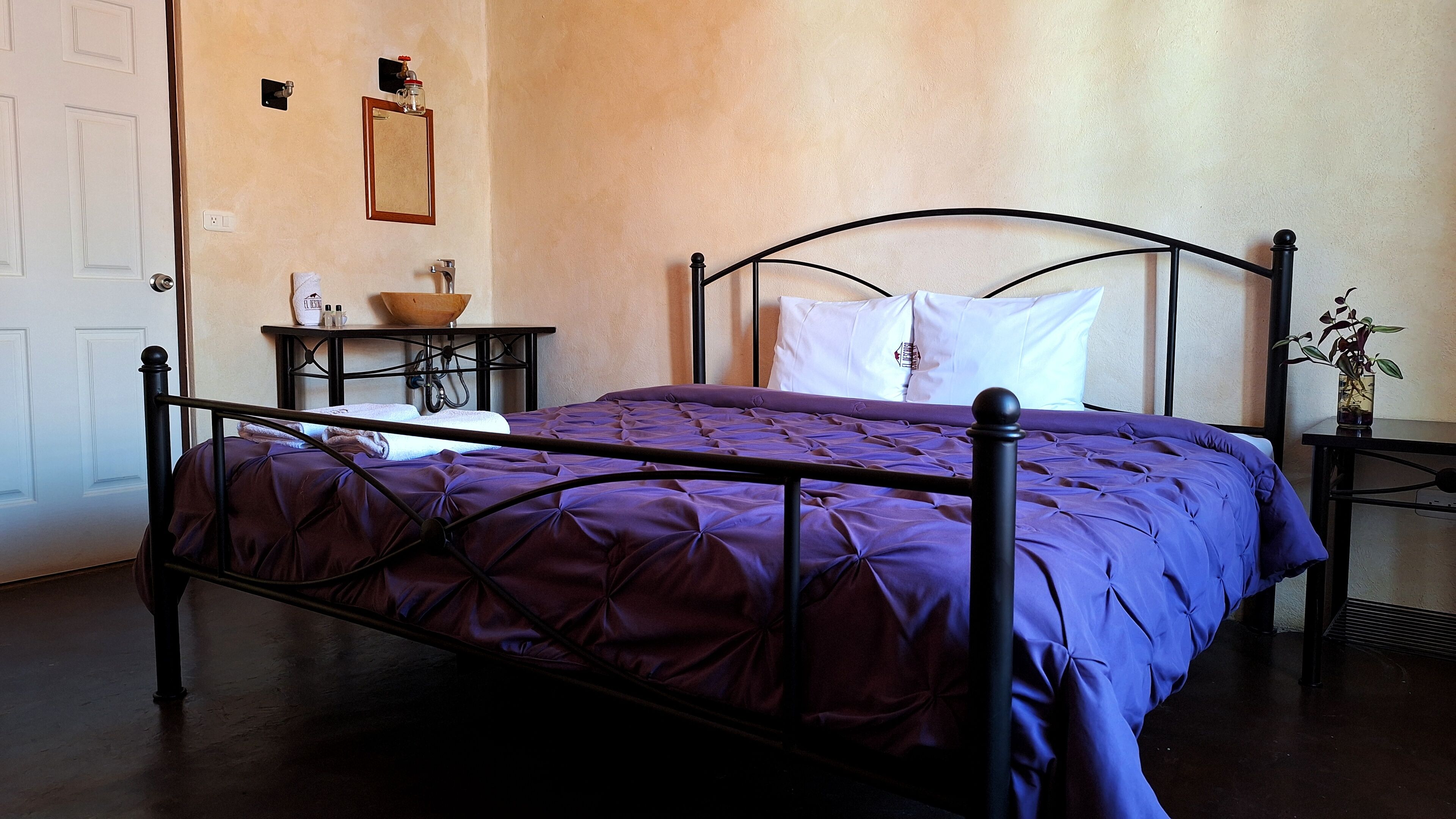 A neatly made bed with a purple bedspread and a metal frame, a small bedside table with a bowl and bottles, and a wall-mounted mirror.