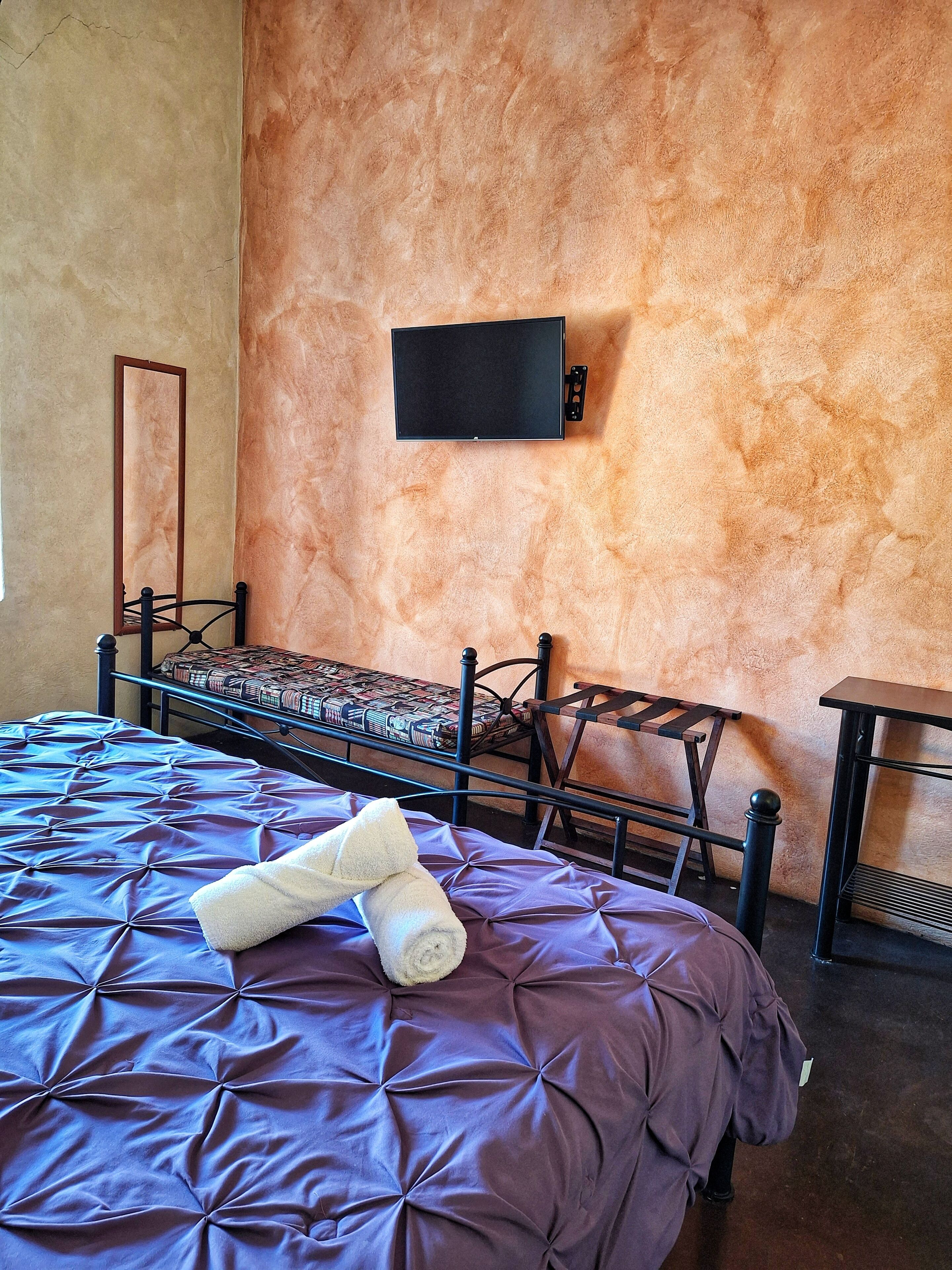 A room with a bed, a chair, a small table, and a flat-screen TV mounted on the wall.