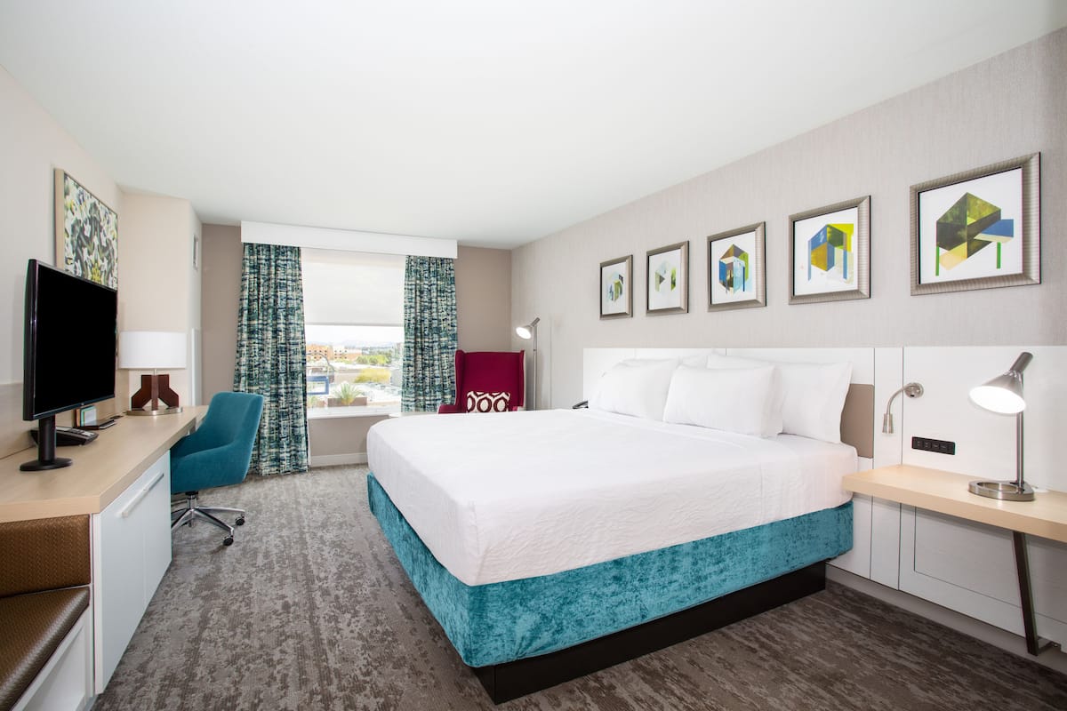A hotel room with a bed, desk, chair, TV, and artwork on the wall.