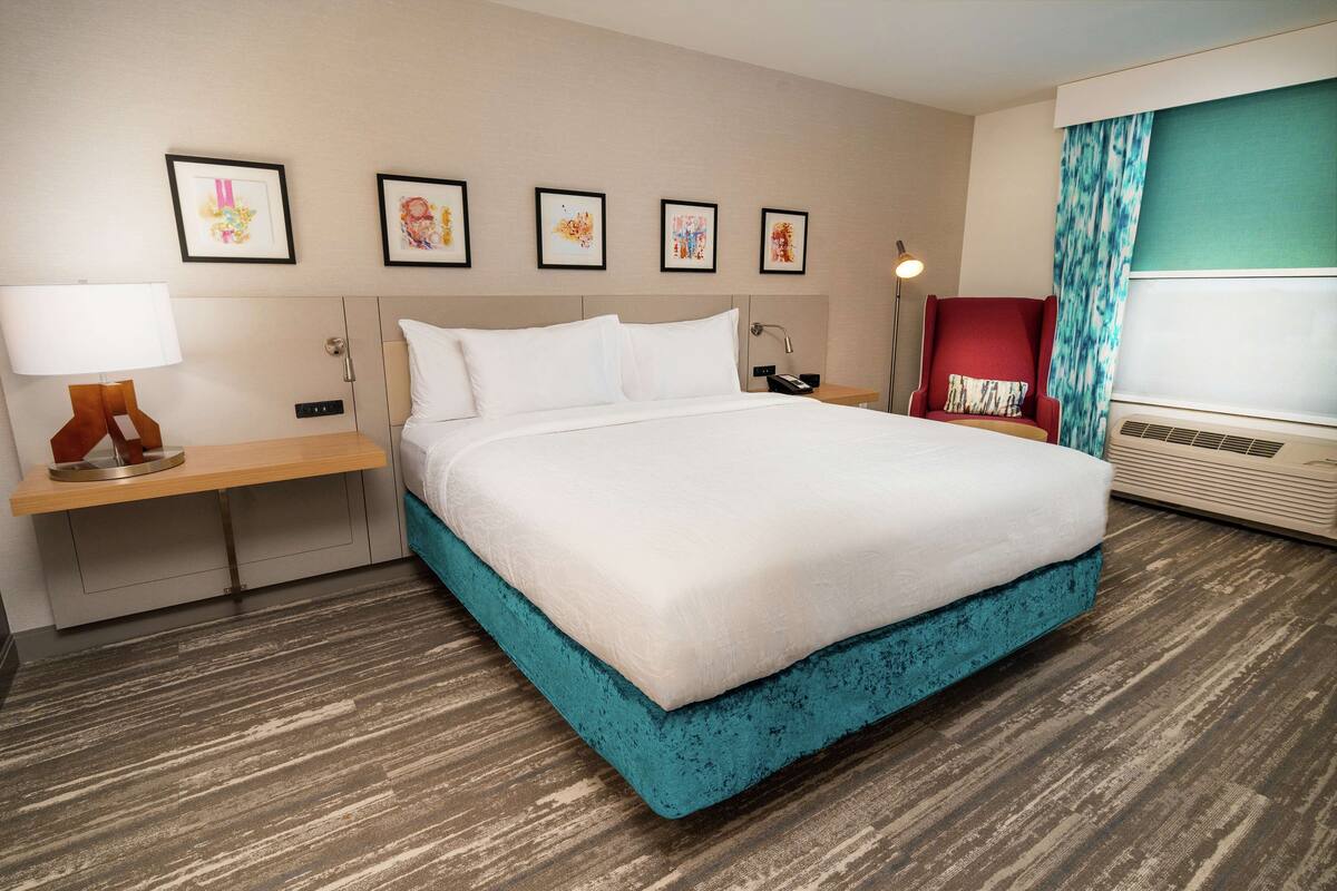 A hotel room with a large bed, a bedside table with a lamp, a red armchair, and a wall with framed pictures.