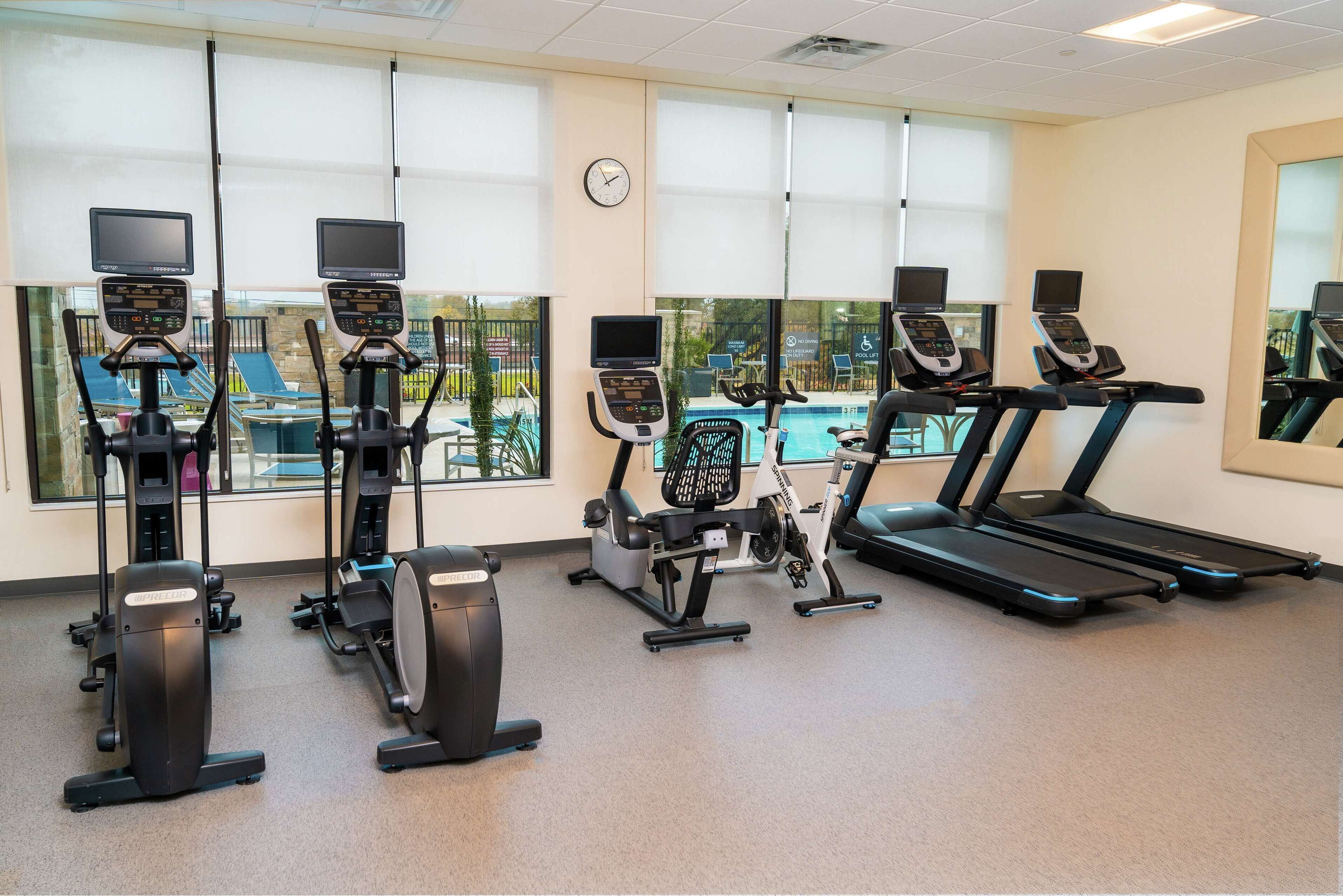 A gym with various exercise machines, large windows, and a clock on the wall.
