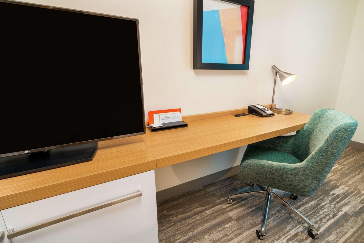A hotel room with a wooden desk, a green upholstered chair, a flat-screen TV, a desk lamp, and a framed abstract painting on the wall.
