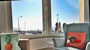 Una habitación con vista a un estacionamiento y a la playa. Hay dos sillas con cojines, una con diseño de piña y la otra con diseño de sandía.