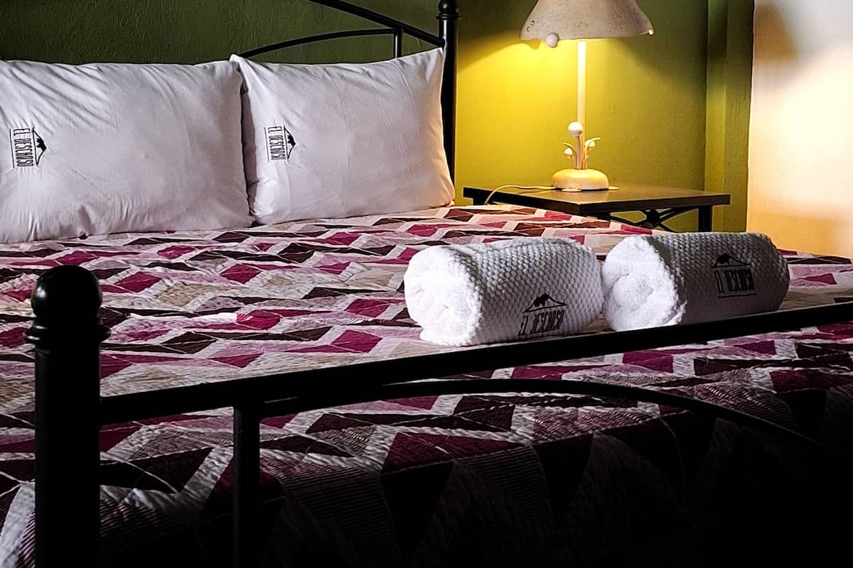 A bed with a quilt, pillows, and rolled towels. There is a lamp on a table beside the bed.