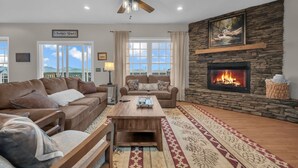 A living room with a stone fireplace, a ceiling fan, and a large window with a view of the mountains. - Blue Ridge View Cabin: Hot Tub, Pool Table, Pet-Friendly, Near Blowing Rock & Parkway (Boone)