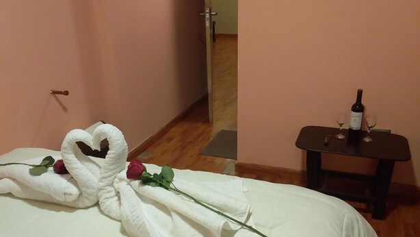 Une chambre d’hôtel avec un lit, une petite table sur laquelle se trouvent une bouteille de vin et des verres, et une porte visible au fond.