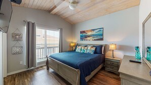 Ein Schlafzimmer mit Holzbalkendecke, einem Bett mit blauer Steppdecke, zwei Wandleuchten, einem Bild eines Strandes und einem Fenster mit Jalousien.