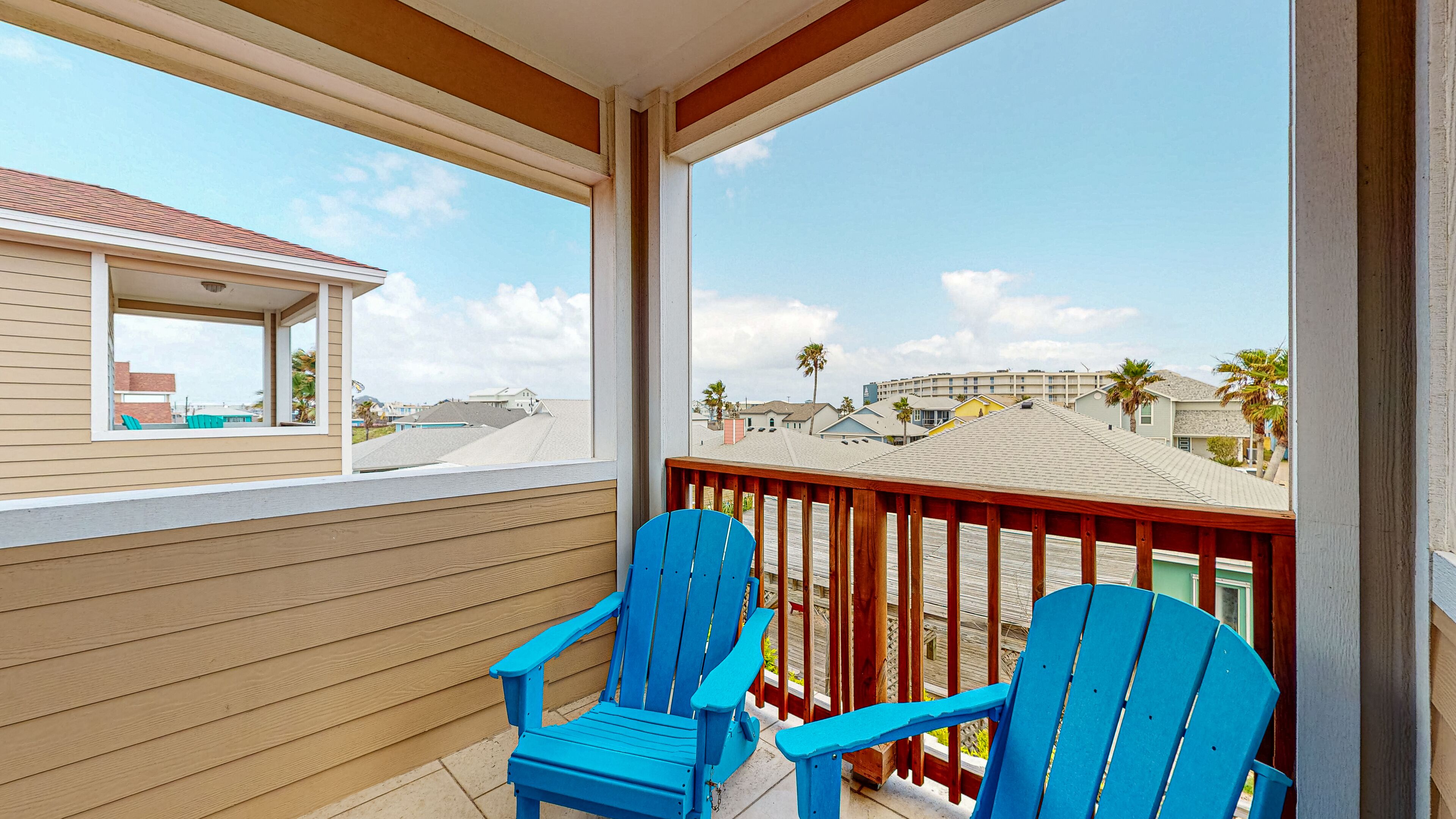 Un balcon avec deux fauteuils Adirondack bleus, une balustrade en bois et une vue sur les maisons voisines et les palmiers.