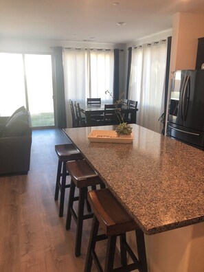 Een moderne keuken met een granieten aanrecht, houten krukken, een eettafel met stoelen en een koelkast.