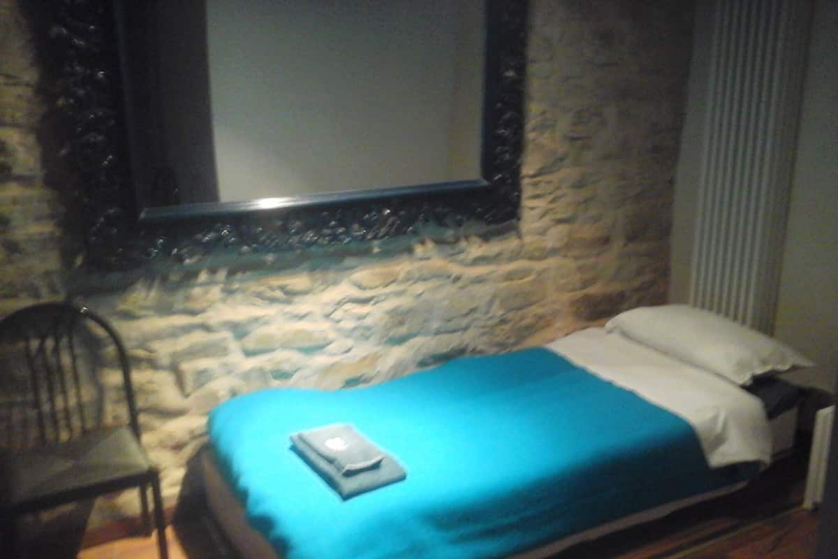 Ein kleines Zimmer mit einem Einzelbett, einer Steinsandsteinwand, einem Stuhl und einem Bett mit blauer Decke und Kissen.