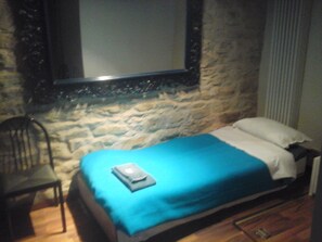 Ein kleines Zimmer mit einem Einzelbett, einer Steinsandsteinwand, einem Stuhl und einem Bett mit blauer Decke und Kissen.