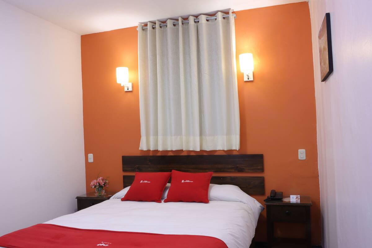 A hotel room with a bed, two red pillows, a nightstand, a vase with flowers, and a window with curtains.