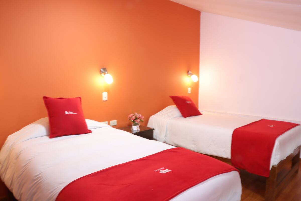 Two single beds with red pillows and white bedding in a room with orange walls and two wall-mounted lights.
