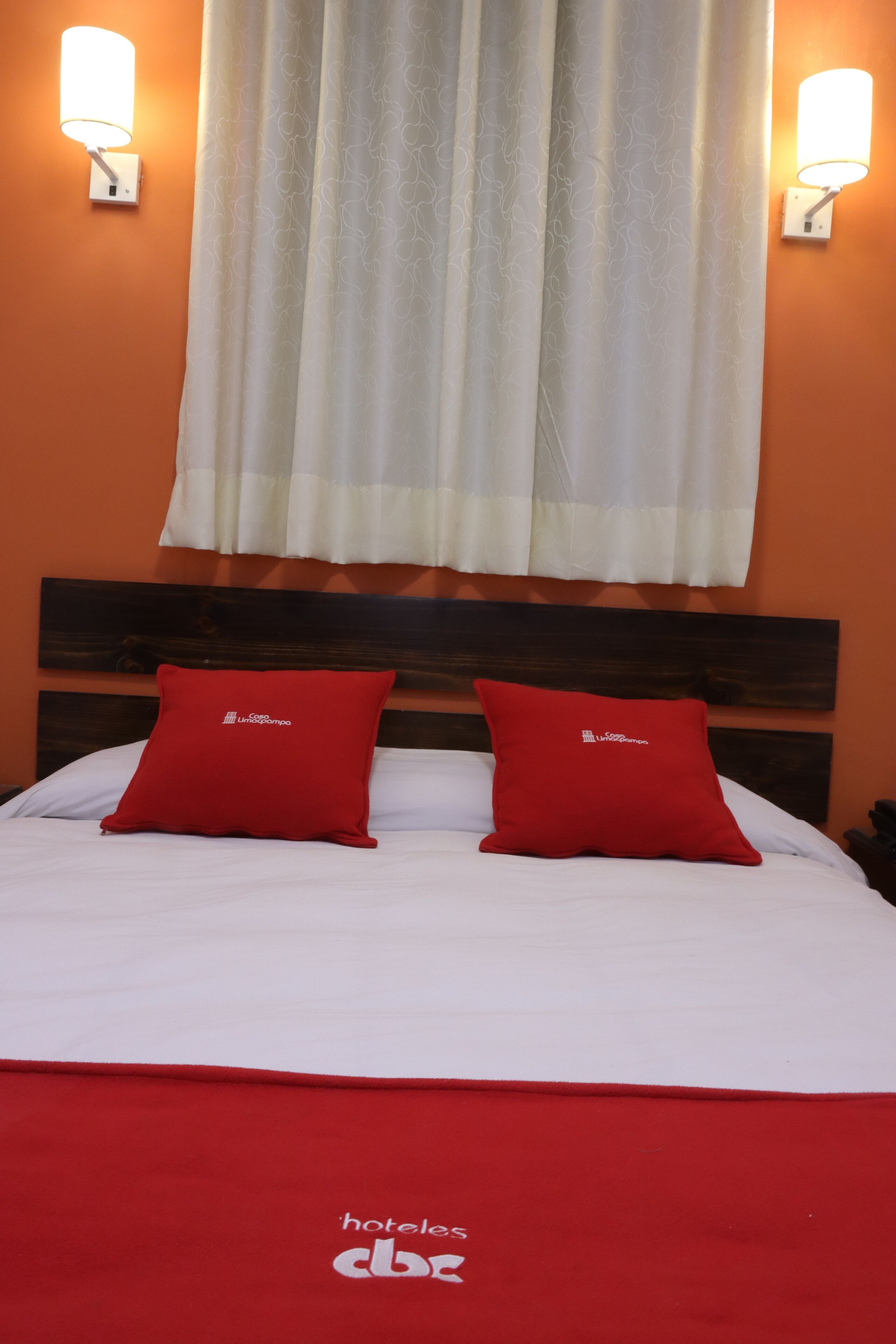 A hotel room with a wooden headboard, two red pillows, a white bedspread, and two wall-mounted lights.