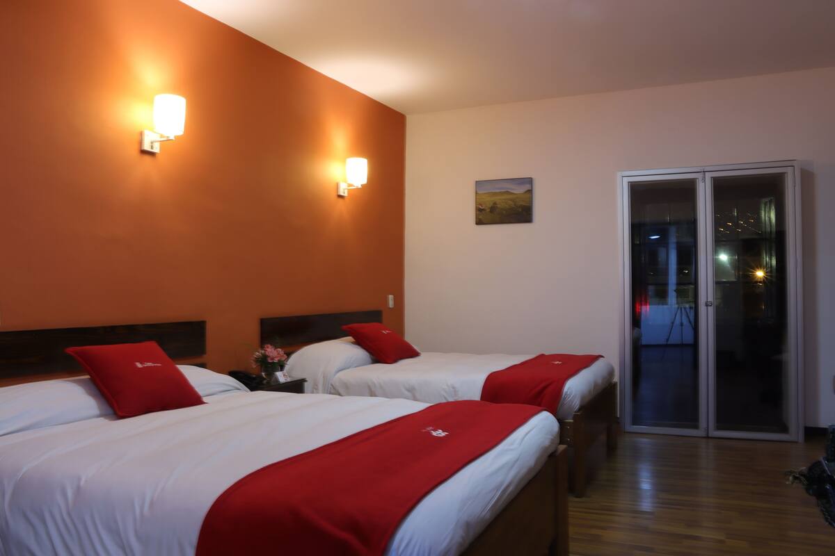 A hotel room with two beds, red pillows, a nightstand with a vase, and a painting on the wall.