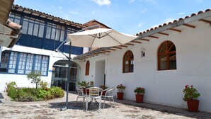 Un patio con una mesa y sillas bajo un paraguas grande, rodeado de plantas en macetas y un edificio con techo de tejas y ventanas arqueadas.